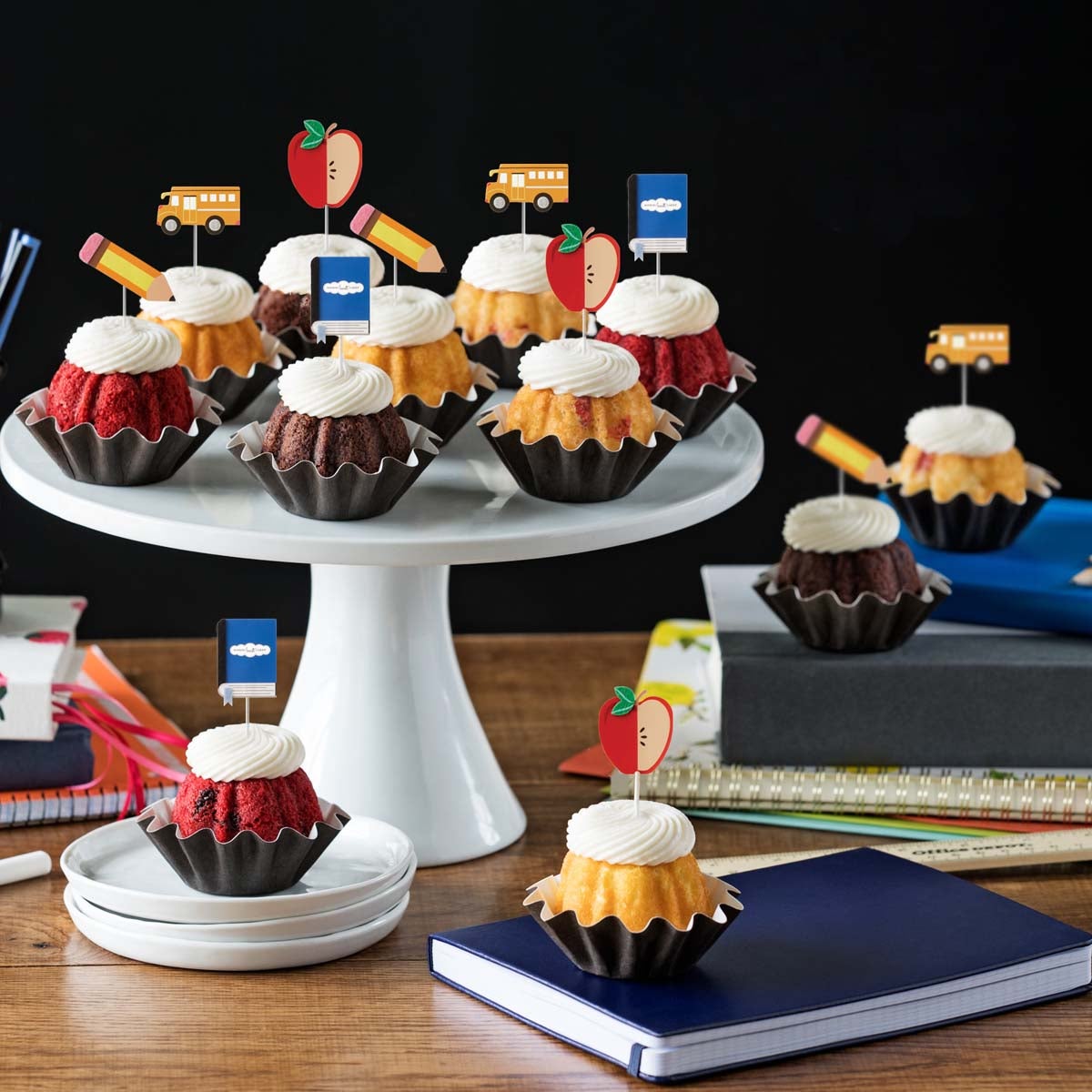 Bundtinis® topped with creamy white frosting, accompanied by school-themed toppers: a sliced apple, a pencil, a notebook, and a yellow school bus.