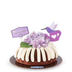 Large cake with frosting on top. The center of the bundt cake is covered with a silk hydrangea flower. There is a hummingbird cutout also decorating the cake and a card that reads best mom a'round.