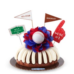 A bundt cake with frosting, decorated with a baseball on top of a purple and white bow in the center of the cake. The cake also contains a We're #1 foam finger, a flag that says All Star and a card that says M.V.P.