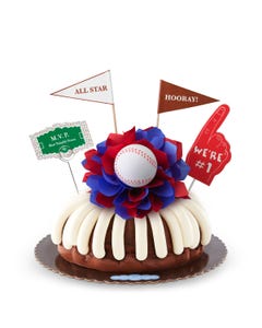 A bundt cake with frosting, decorated with a baseball on top of a purple and white bow in the center of the cake. The cake also contains a We're #1 foam finger, a flag that says All Star and a card that says M.V.P.