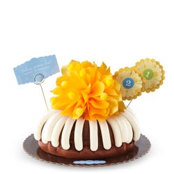 Birthday bundt cake with frosting all around. Topped with a golden yellow bow and numbered flowers suitable for age or any numbered milestone. 