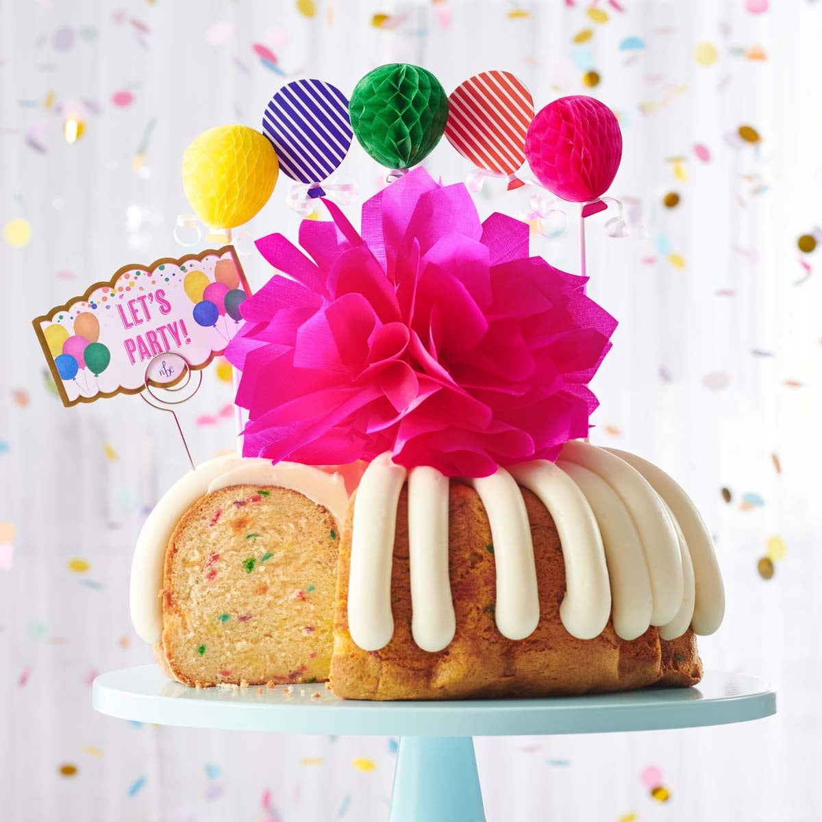 A party-themed bundt cake with colorful 3D balloons. The cake has a card that reads LET'S PARTY. 