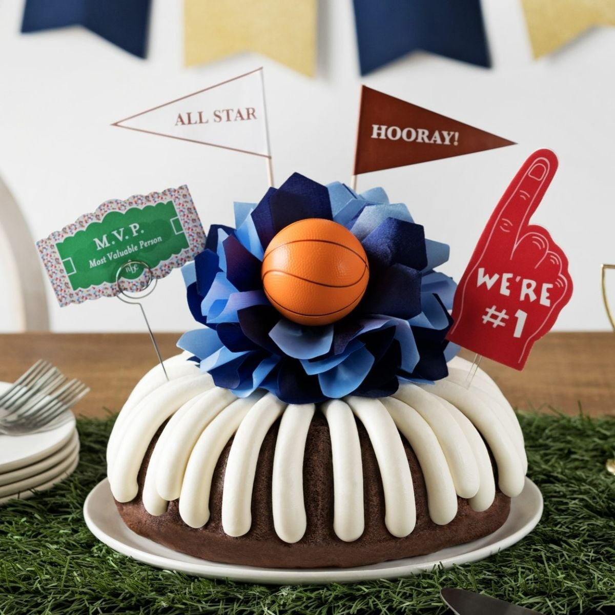 A bundt cake with frosting, decorated with a mini basketball on top of a bow with different shades of blue in the center of the cake. The cake also contains a We're #1 foam finger, a flag that says All Star and a card that says MVP.