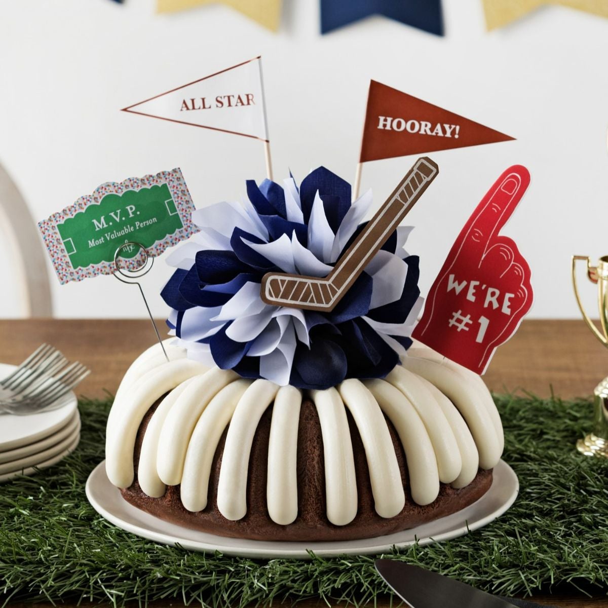 A bundt cake with frosting, decorated with a mini hockey puck on top of a blue and white bow in the center of the cake. The cake also contains a We're #1 foam finger, a flag that says All Star and a card that says M.V.P.