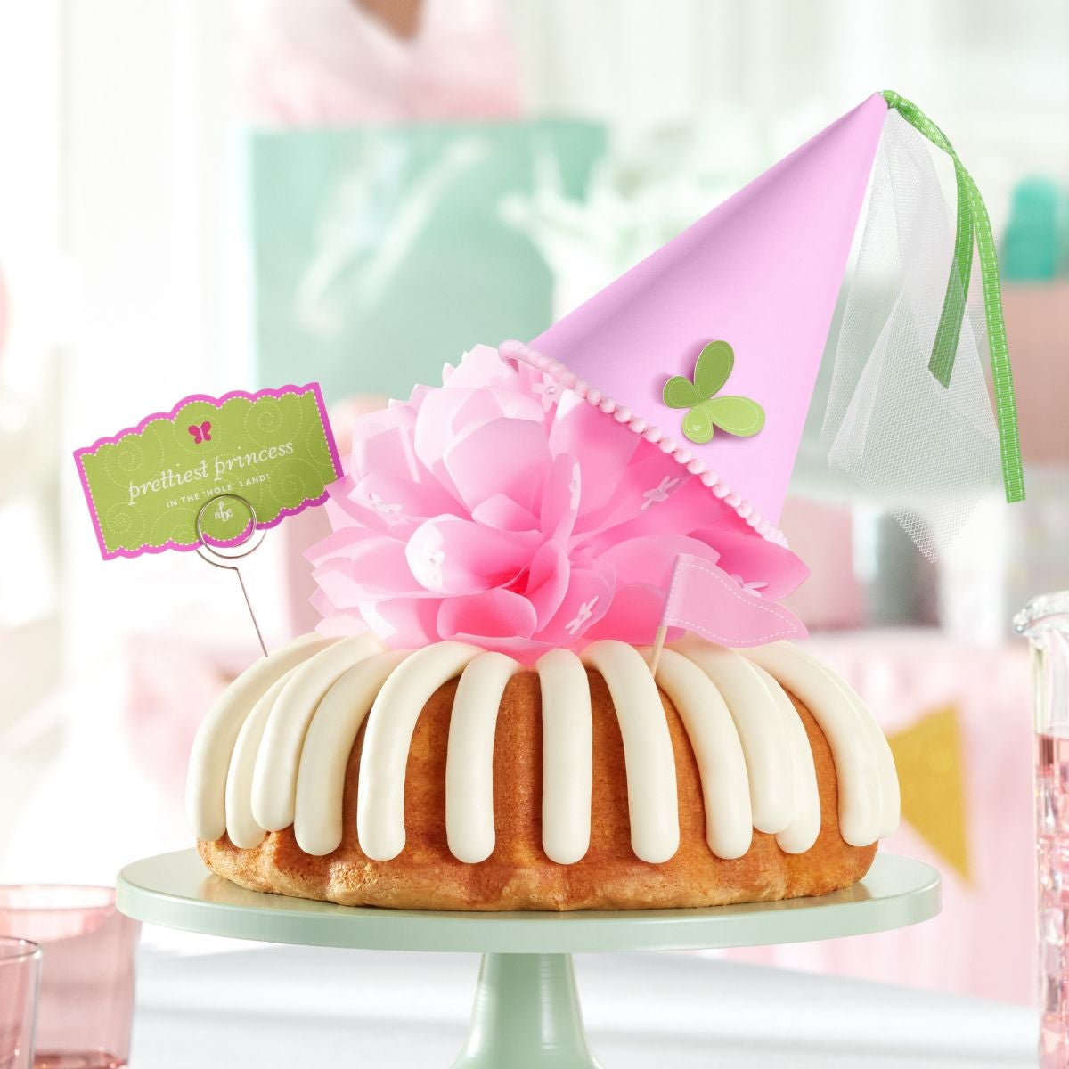 A bundt cake with frosting, topped with a pink bow and a princess-themed pink party hat with a butterfly on top. A card on the cake reads prettiest princess. 