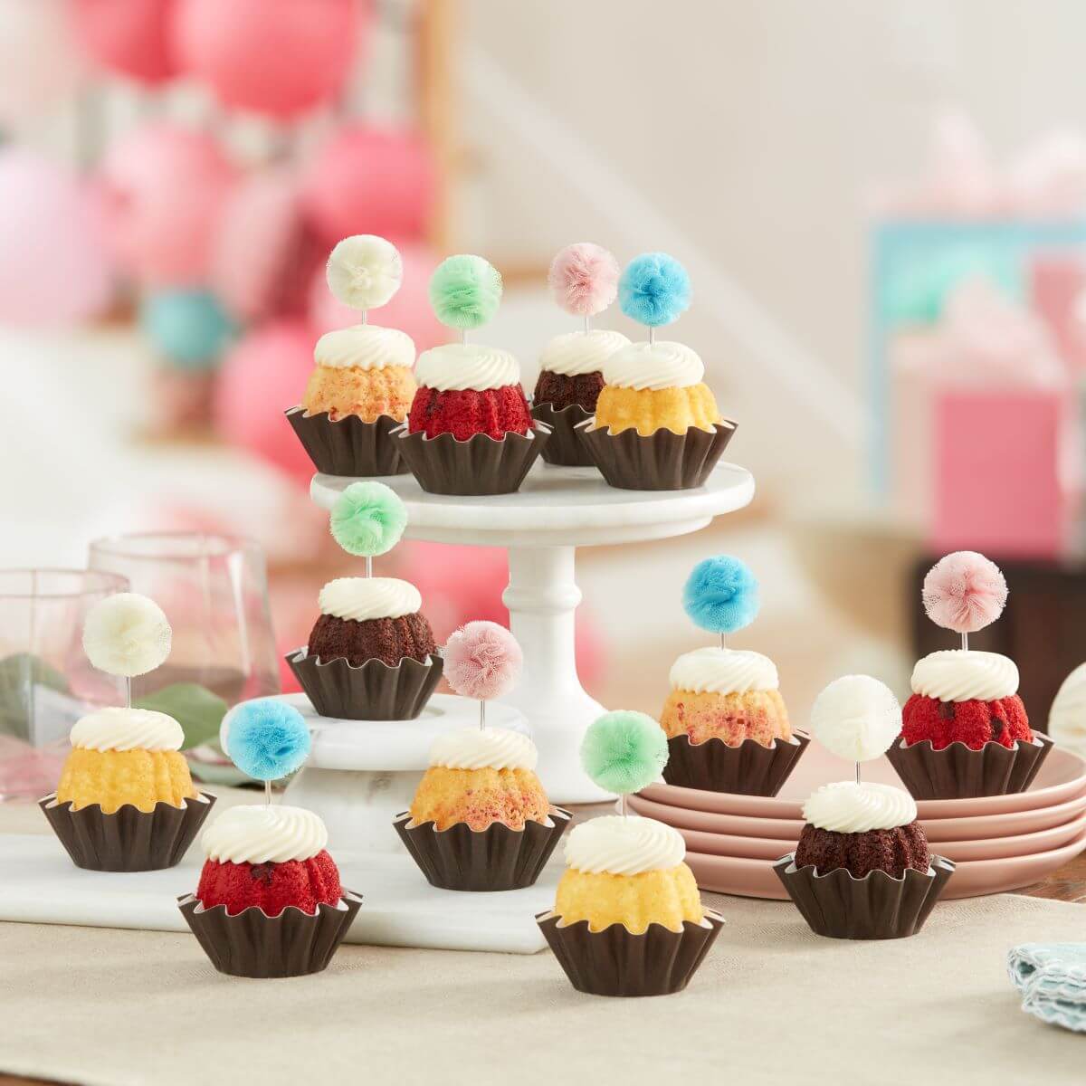 Cupcake-sized Bundt cakes topped with creamy white frosting. Each has a fluffy pom-pom topper in shades of sky blue, white, mint green, and soft rose.