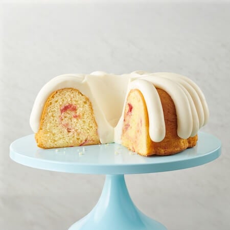 White chocolate strawberry Bundt cake on a cake stand, a slice cut out showing the middle of the cake with - Shop Bundt Cakes